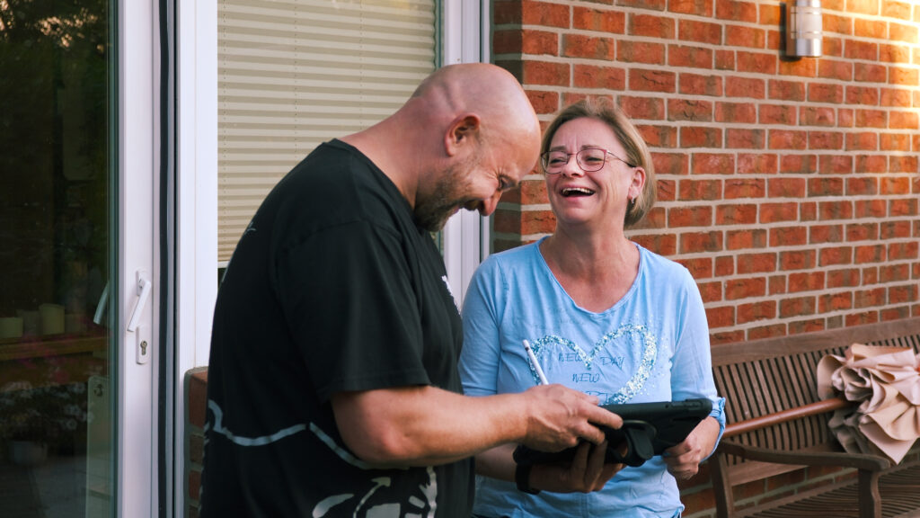 Techniker und Kundin stehen lachend vor einer Hauswand aus rotem Klinker und schauen gemeinsam auf ein Tablet.