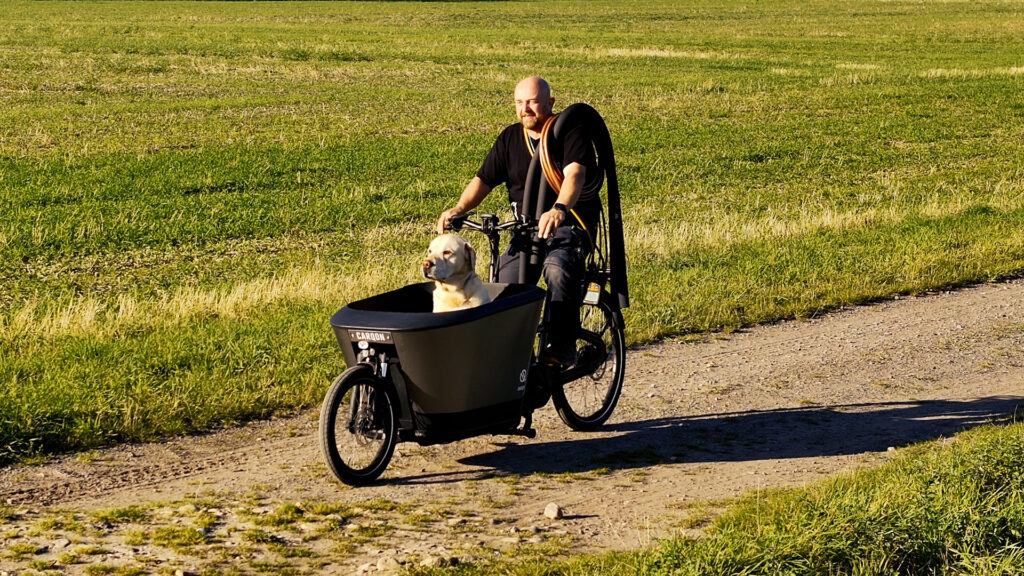 Mann fährt auf einem Lastenrad über einen Feldweg, im vorderen Transportbehälter sitzt ein Hund.