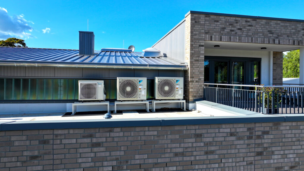 Drei Außengeräte einer Klimaanlage auf einem Flachdach vor einem modernen Gebäude mit Klinkerfassade und Metallgeländer.