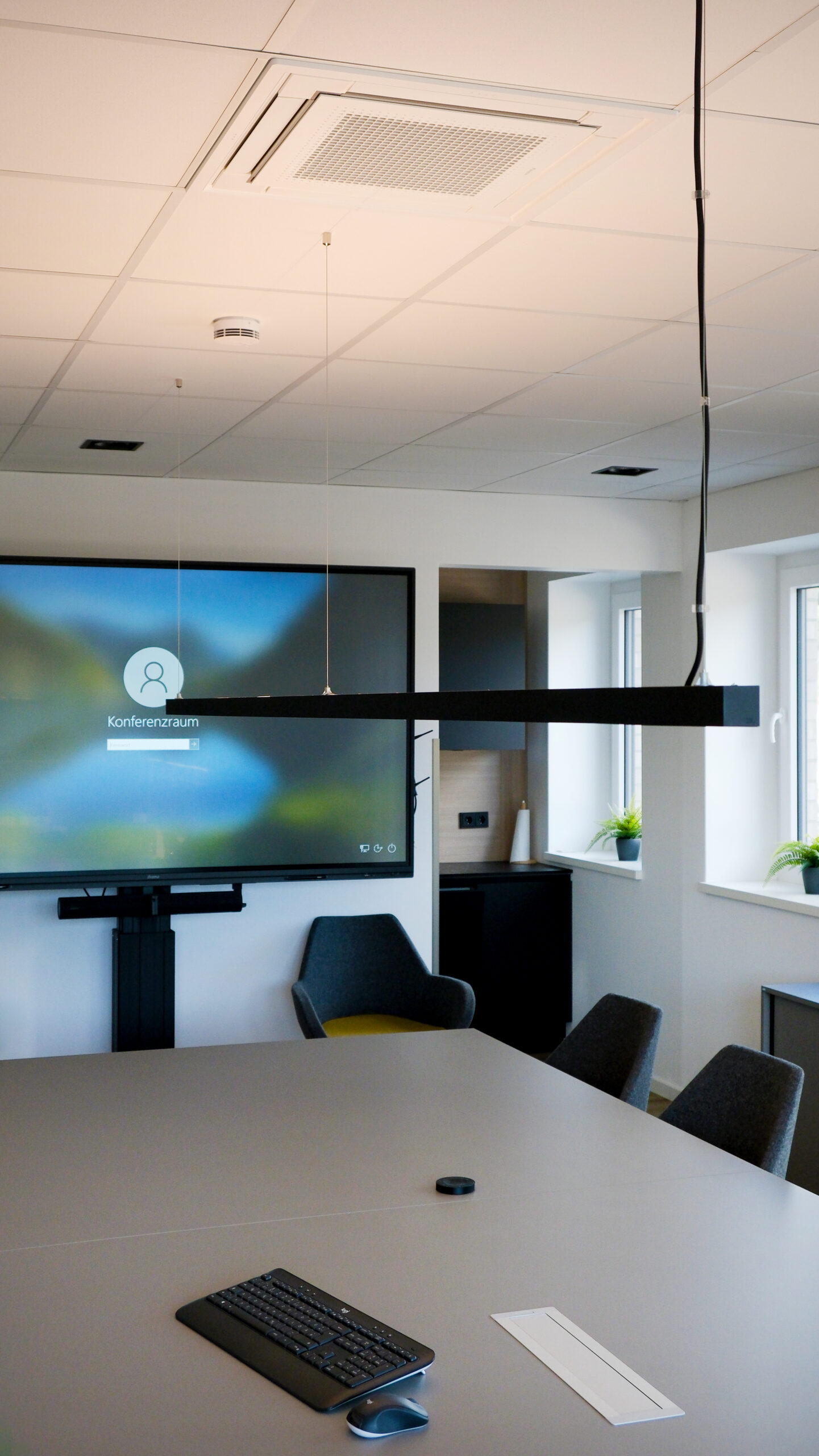 Moderner Konferenzraum mit Deckenklimaanlage, großem Bildschirm, Besprechungstisch mit Tastatur und grauen Stühlen.