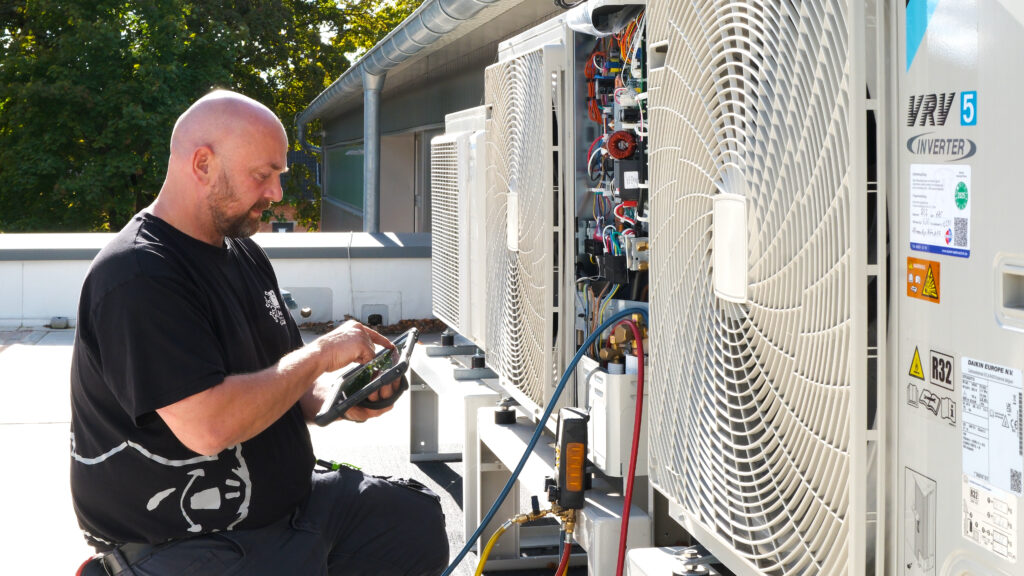 Techniker prüft eine Klimaanlage auf dem Dach und liest Messwerte auf einem Tablet ab.