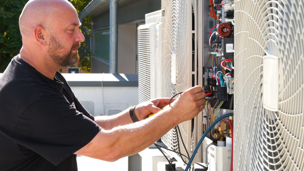 Techniker arbeitet an geöffnetem Außengerät einer Klimaanlage und misst elektrische Anschlüsse mit einem Messgerät.
