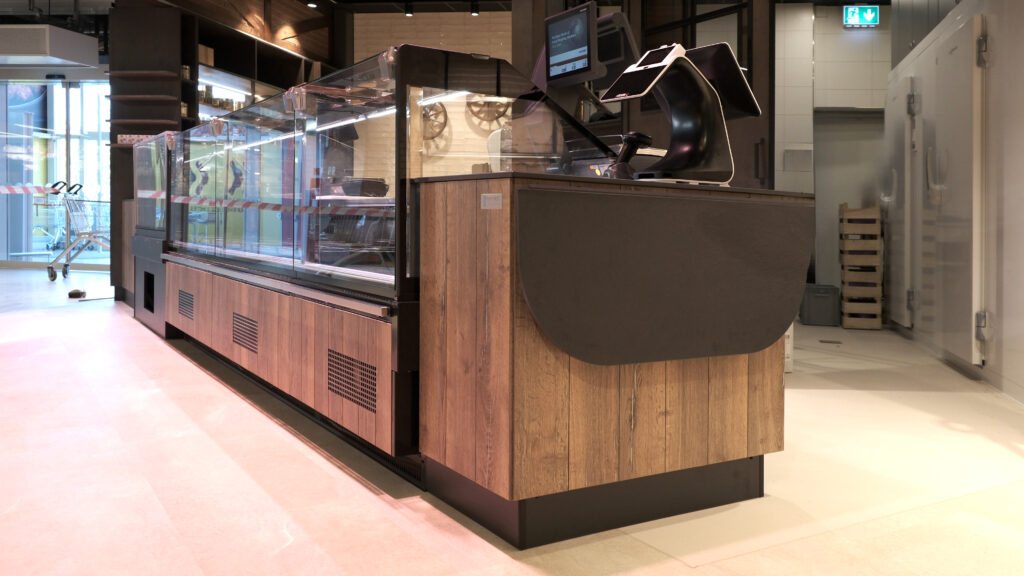 Moderne Frischetheke mit Holzverkleidung und Glasvitrine in einem Lebensmittelmarkt, daneben Kassenmonitor und Kühlraumtüren im Hintergrund.