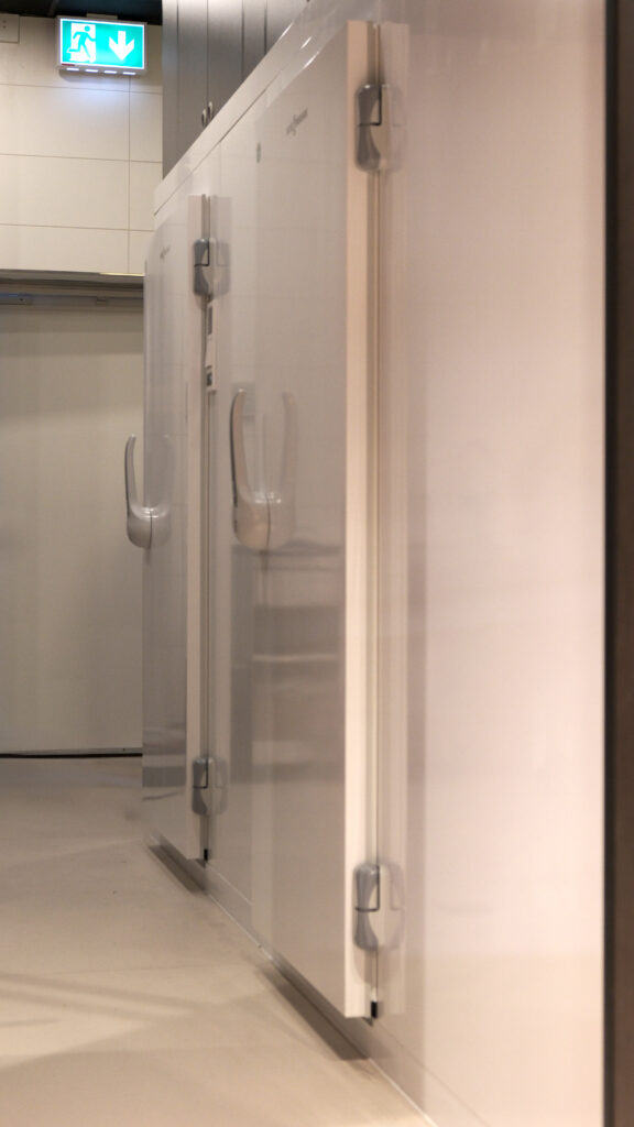Weiße Kühlzellen mit Metallgriffen in einem Flur, darüber ein leuchtendes grünes Notausgangsschild