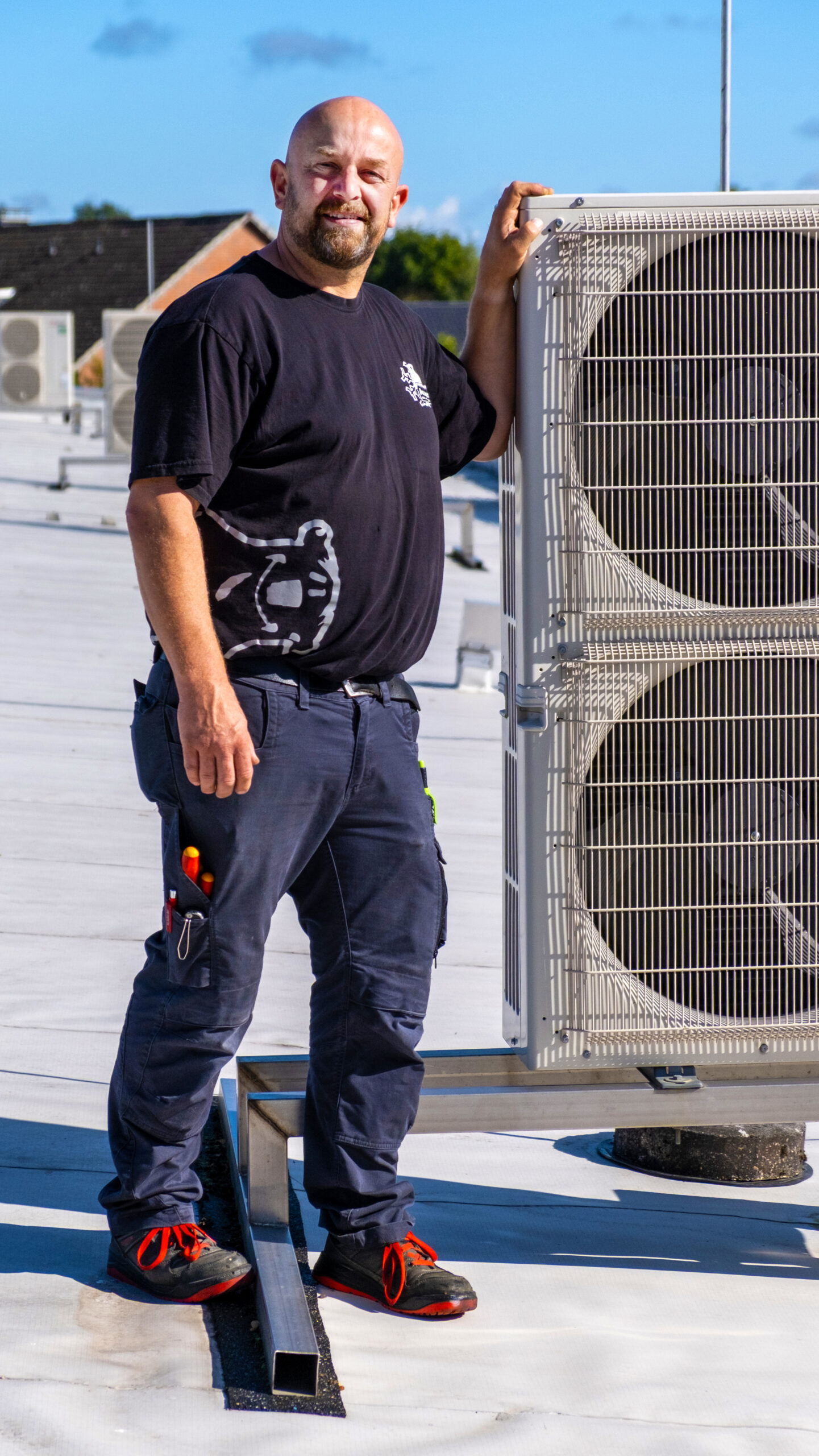 Handwerker mit Werkzeugen in der Hosentasche steht auf einem Flachdach neben einer großen Klimaanlagen-Außeneinheit.