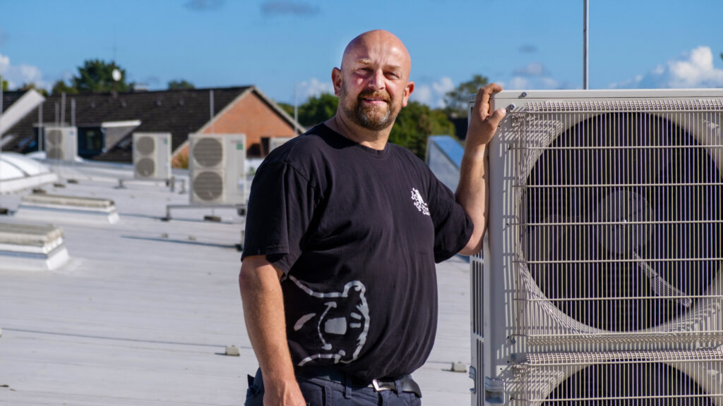 Techniker steht auf einem Flachdach neben einer großen Klimaanlagen-Außeneinheit und blickt in die Kamera