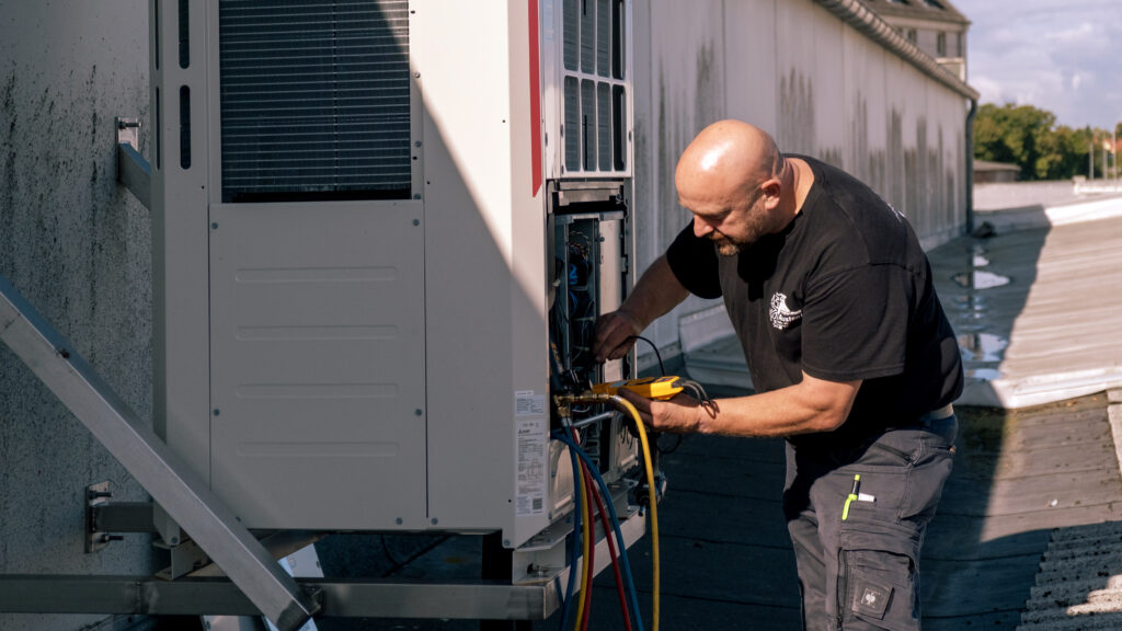 Techniker arbeitet mit Messgerät und farbigen Schläuchen an einer geöffneten Klimaanlagen-Außeneinheit auf einem Flachdach.