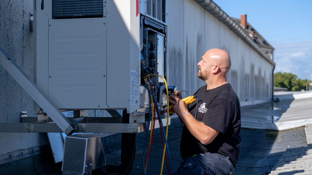 Techniker überprüft auf einem Flachdach eine geöffnete Außeneinheit einer Klimaanlage und misst Werte mit einem Diagnosegerät