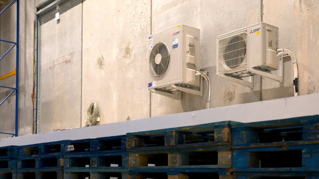 Zwei Mitsubishi-Electric Außengeräte für Klimaanlagen an einer Betonwand in einer Industriehalle, davor gestapelte blaue Holzpaletten