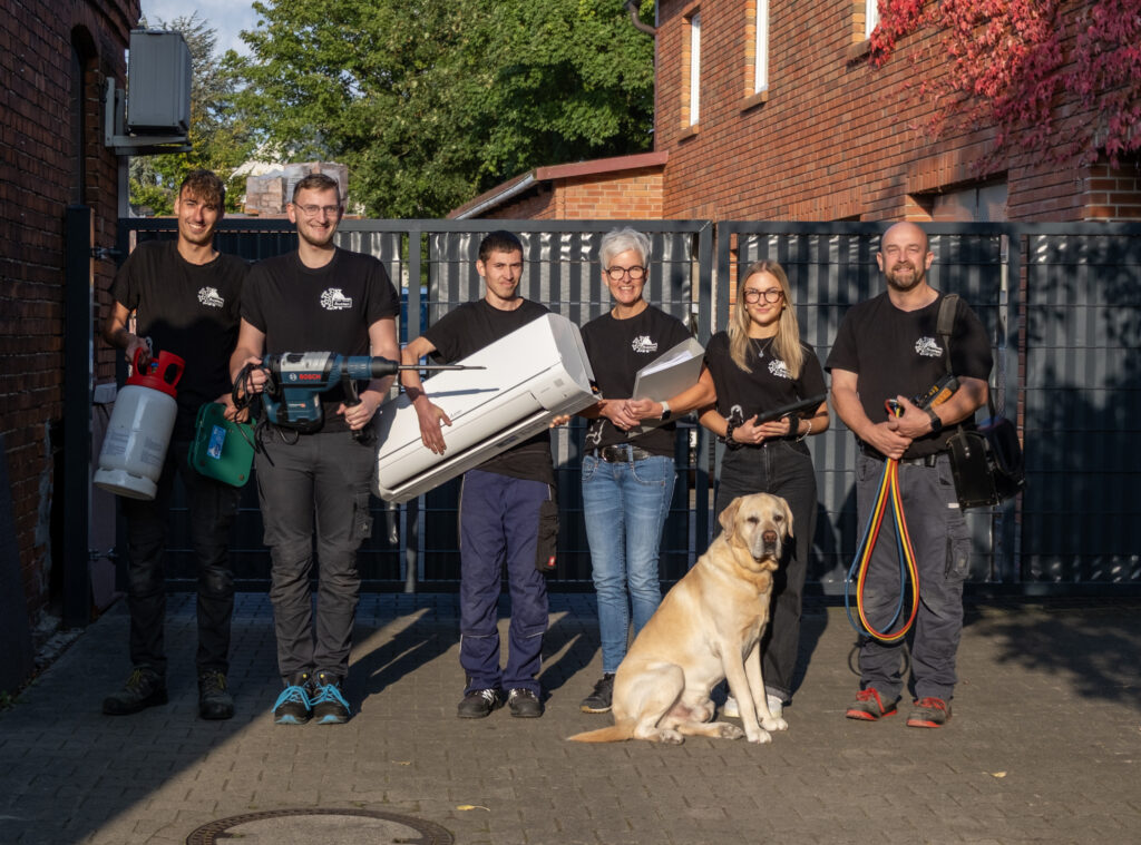 Team der Austen Kälte-Klima GmbH mit Werkzeugen und Klimagerät, stehend vor dem Firmenhof, begleitet von einem hellen Labrador.